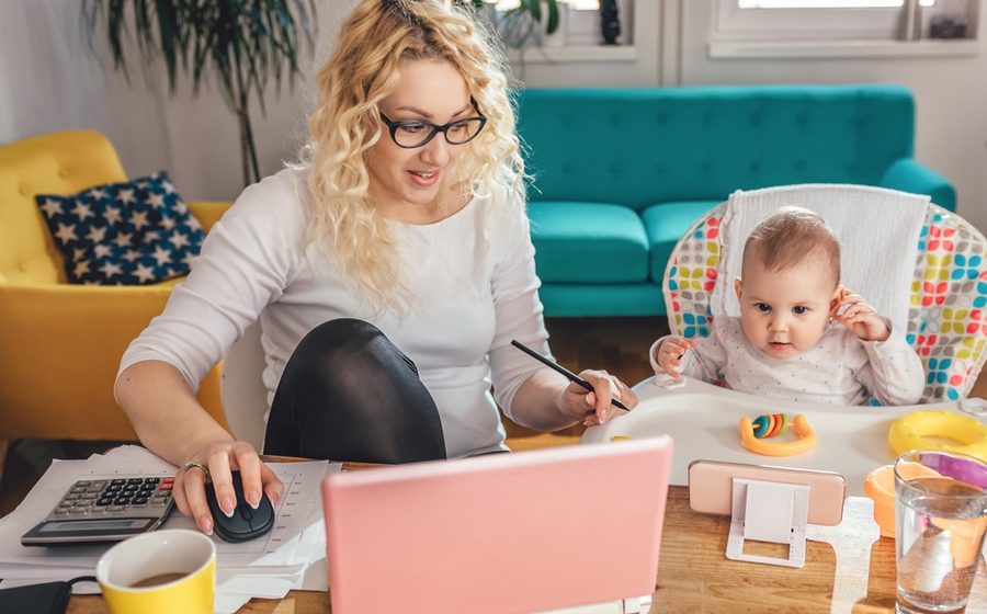 Mutter arbeitet am Laptop, während ihr Baby im Kinderhochstuhl daneben sitzt